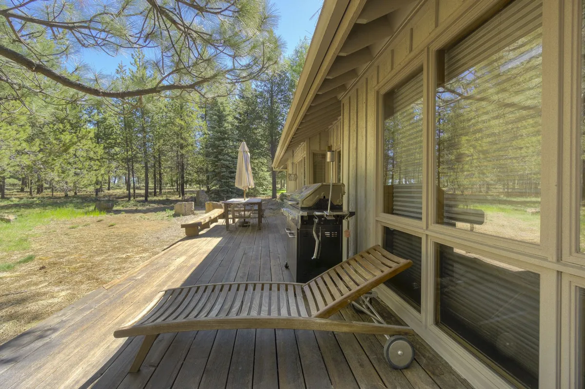 This image features a spacious outdoor deck complete with a grill, dining table, umbrella, and lounge chair, all surrounded by lush trees. The deck invites gatherings for meals, relaxation, and enjoying the natural setting, capturing the essence of indoor-outdoor living.