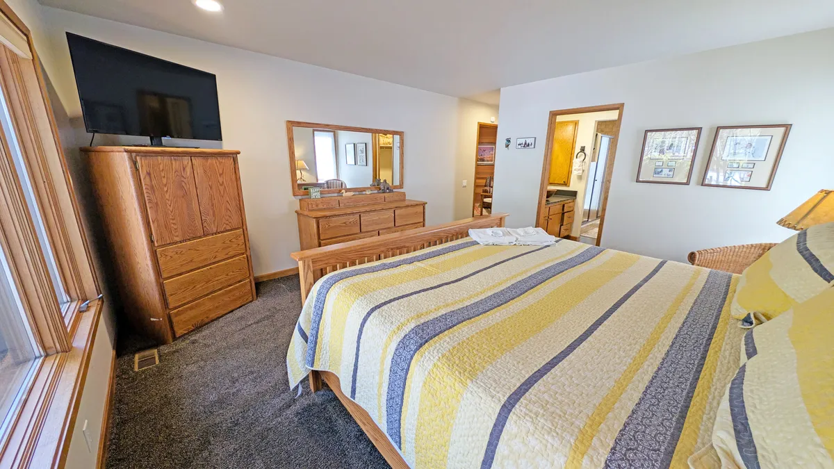 A tranquil bedroom with a bed dressed in a yellow and gray striped quilt, complemented by wooden furniture including a dresser and chest. A large window fills the room with natural light, creating an inviting and restful atmosphere.