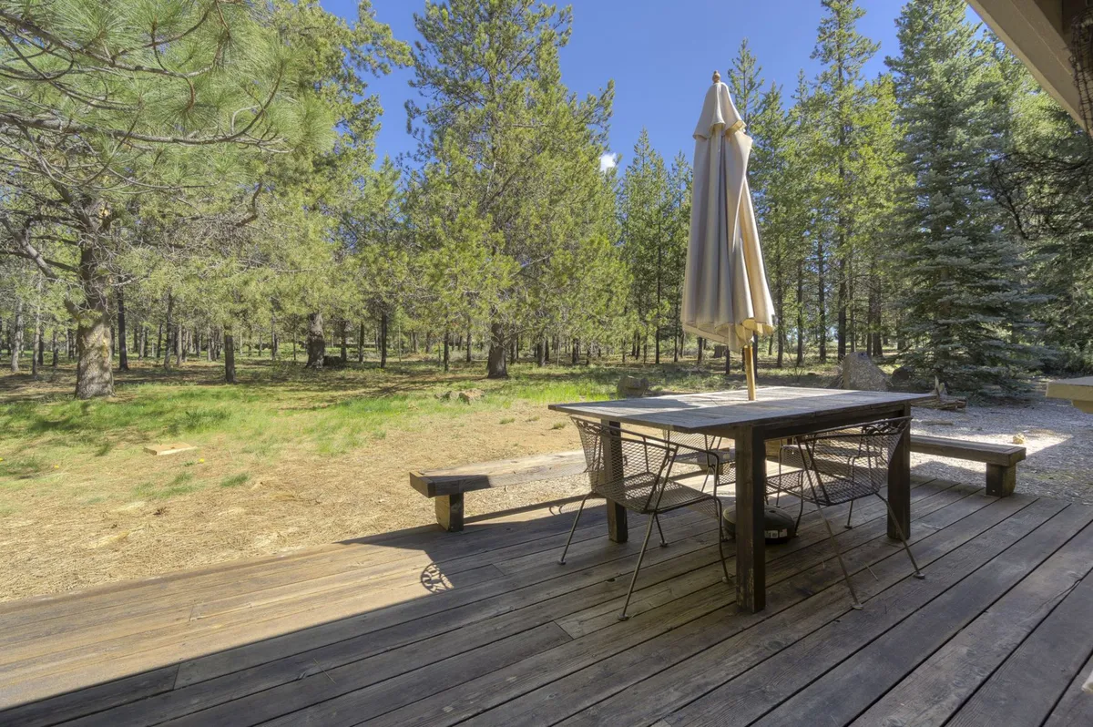 A wooden deck with an outdoor dining table and parasol, surrounded by tall trees. The setup is inviting for alfresco dining, casual gatherings, and relaxation with nature as a backdrop.
