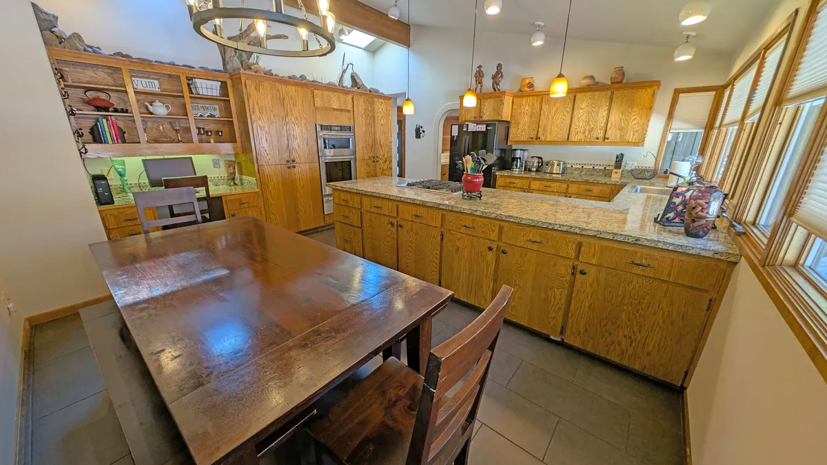 An open-concept area with a large wooden dining table, granite kitchen island, open shelving adorned with decorative pieces, and stylish hanging light fixtures. The space is filled with natural light, perfect for family meals and entertaining.