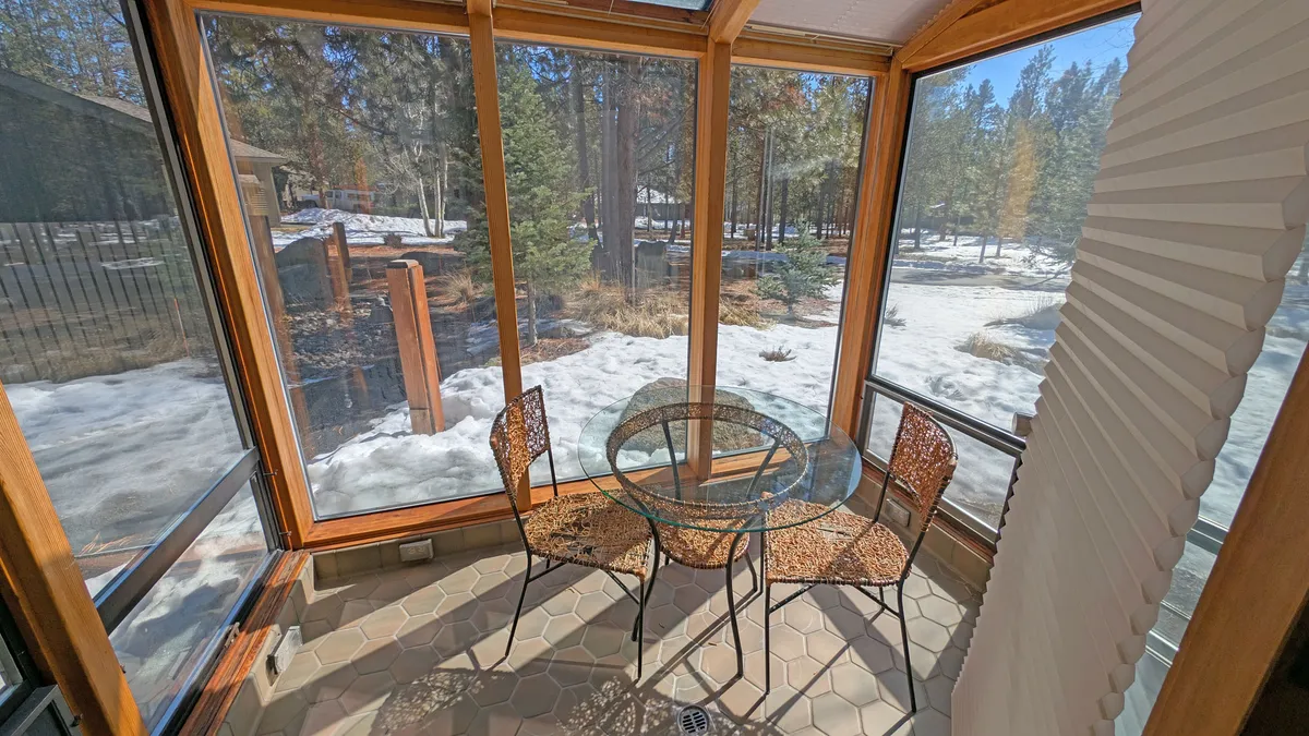 A sunny glass-walled sunroom with a glass table and woven chairs, surrounded by lush nature views outside. The space is ideal for morning coffee, reading, or quiet conversation, bathed in the warmth of the sun.
