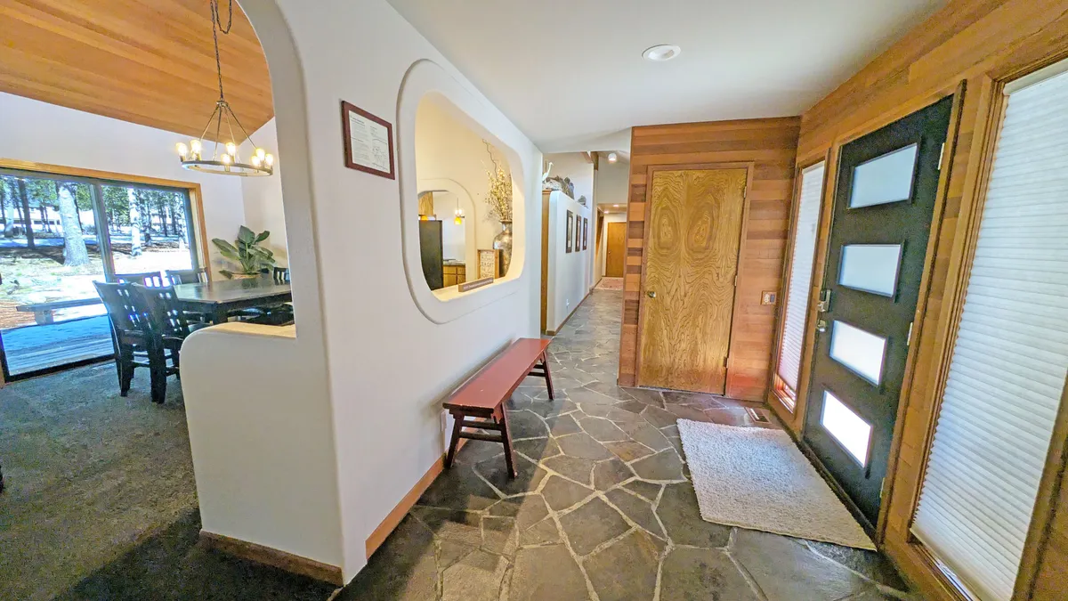 This image shows the home's front door, stone flooring, an arched mirror, and a wooden bench. The dining area is visible through the doorway, and large windows offer hints of natural views outside. The setup creates an inviting transition between interior and exterior spaces.