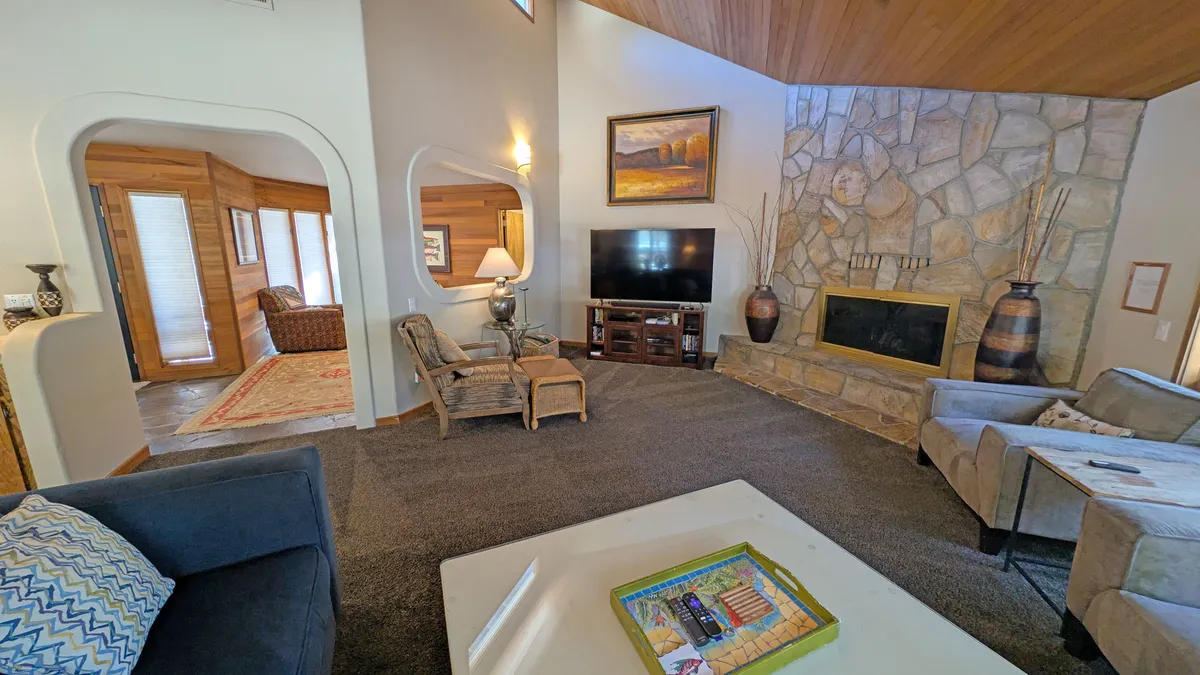 A warm, inviting living room featuring a stone fireplace, a large TV, plush seating arranged for conversation, decorative art, and exposed wooden beams overhead. The space exudes comfort and encourages group relaxation and socializing.