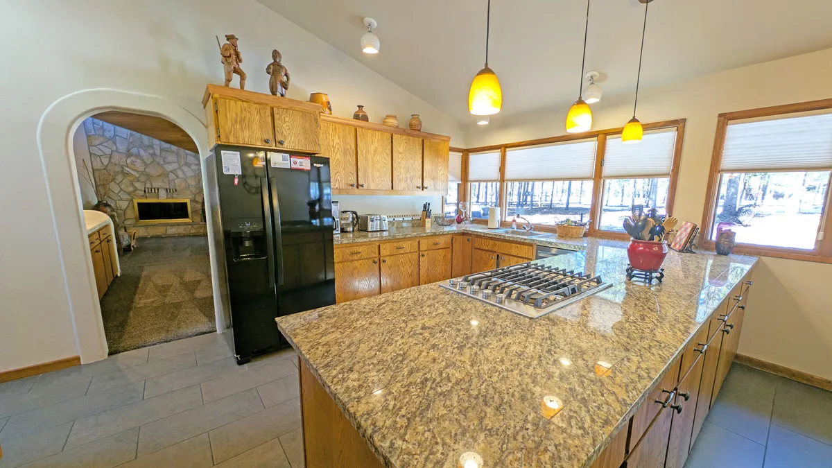 A spacious kitchen with a central island topped with granite, large windows bringing in sunlight, wooden cabinetry, and decorative figures. The layout is ideal for cooking, baking, and hosting, with a welcoming, family-friendly atmosphere.