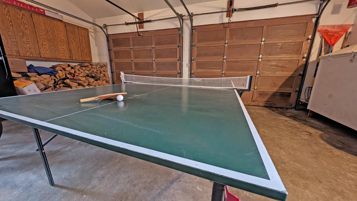 A garage converted into a lively game area, centered around a ping pong table. The scene includes ping pong paddles and balls on the table, with natural light filtering through garage doors and a stack of wood nearby, adding to the casual, active vibe.