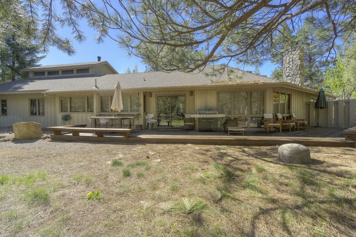 A view of the home's patio area featuring outdoor seating, a grill for barbecuing, a stone chimney, and large windows that blur the line between indoor and outdoor living. The space is surrounded by tall trees, creating a relaxed and social environment.