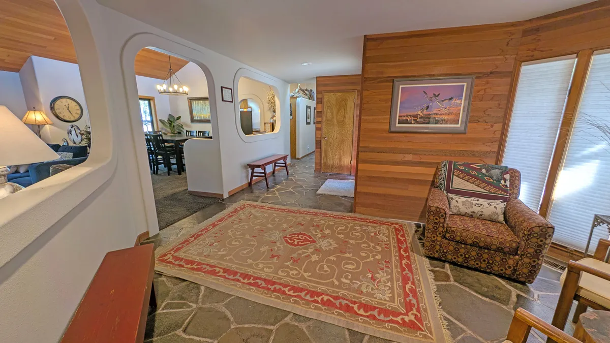 A cozy, light-filled room with a patterned area rug, comfortable chair, wooden door, and arched entryways. The space is set for relaxing, social gatherings, or simply enjoying the view through sunlit windows.
