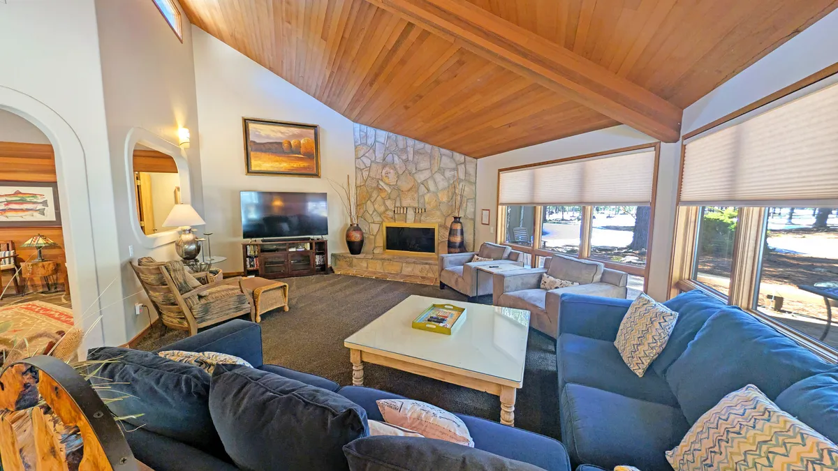 The image showcases a spacious living room with a stunning stone fireplace as the centerpiece, large windows that flood the space with natural light, plush and inviting seating arranged for family gatherings, and a warm wooden ceiling that enhances the cozy mountain-lodge ambiance. The setting is perfect for relaxing, enjoying fireside conversations, or unwinding with a good book or movie after a day of adventure in Sunriver.