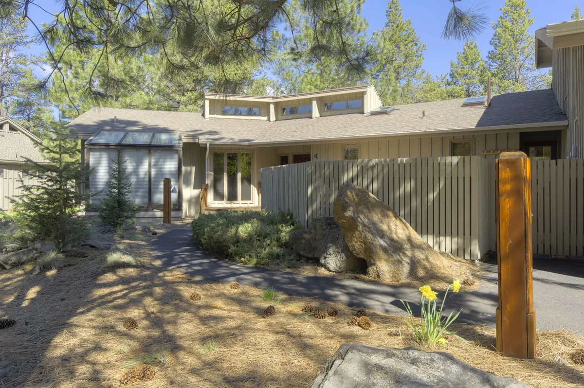 This exterior image shows a tranquil garden pathway leading to the property's wooden entrance, surrounded by natural landscaping and mature trees. The large rock in the foreground and wooden posts reinforce the natural, welcoming atmosphere that guests encounter upon arrival.