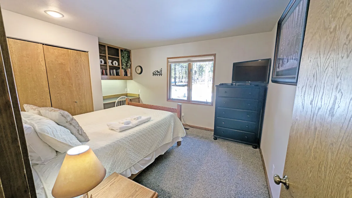 A peaceful bedroom featuring a bed with crisp white linens, a window offering outdoor views, a dresser, and a desk for productivity. The room is filled with soft, natural light, emphasizing its dual function as both a restful and practical space.