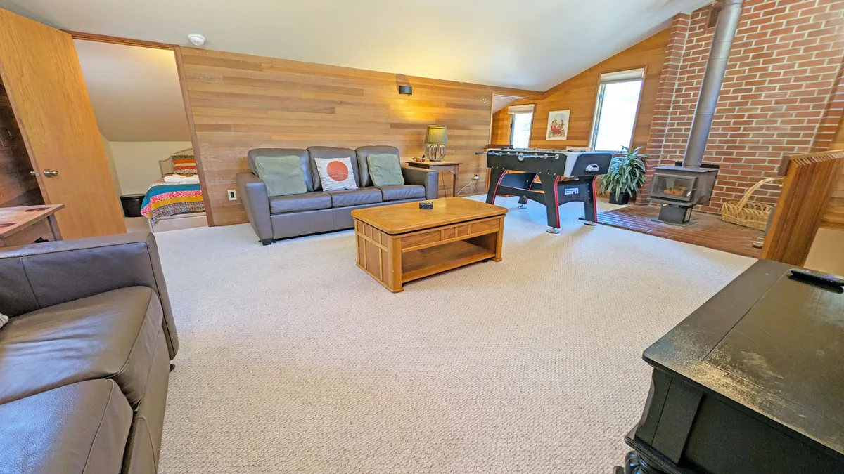 A cozy upstairs loft with a sofa set arranged around a wooden coffee table. The foosball table and wood stove are central, creating a gathering space for games and conversation. Light from nearby windows gives the room a warm, inviting atmosphere.